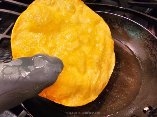 A crispy fried tortilla is removed from skillet using tongs.