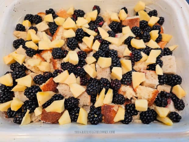 Blackberries and apple pieces are spread on top of bread cubes in dish.