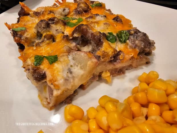 A piece of cheesy beef potato casserole on white plate with corn on the side.